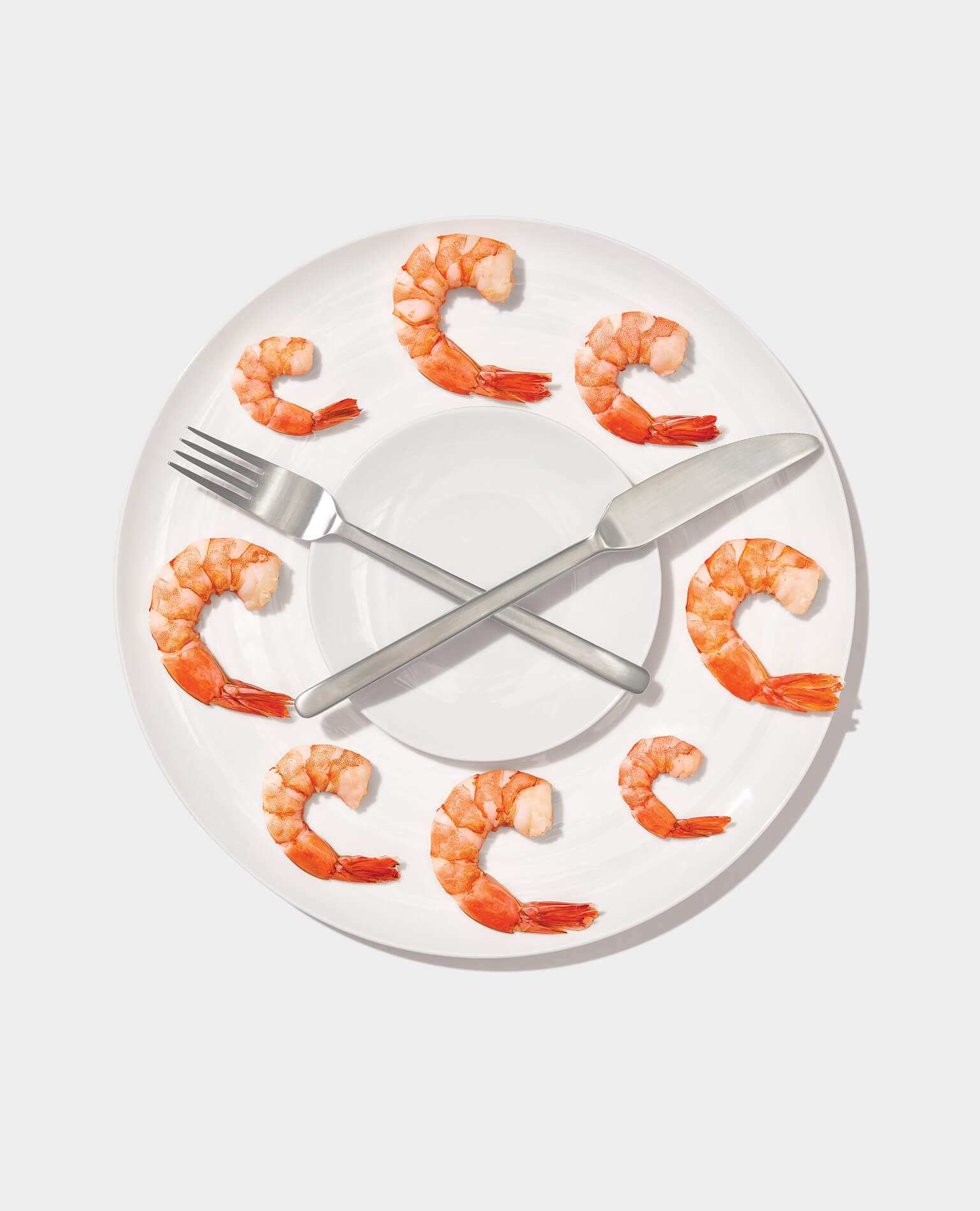 Uncooked shrimp around a plate like the numbers on a clock with a fork and knife as the clock's hands.