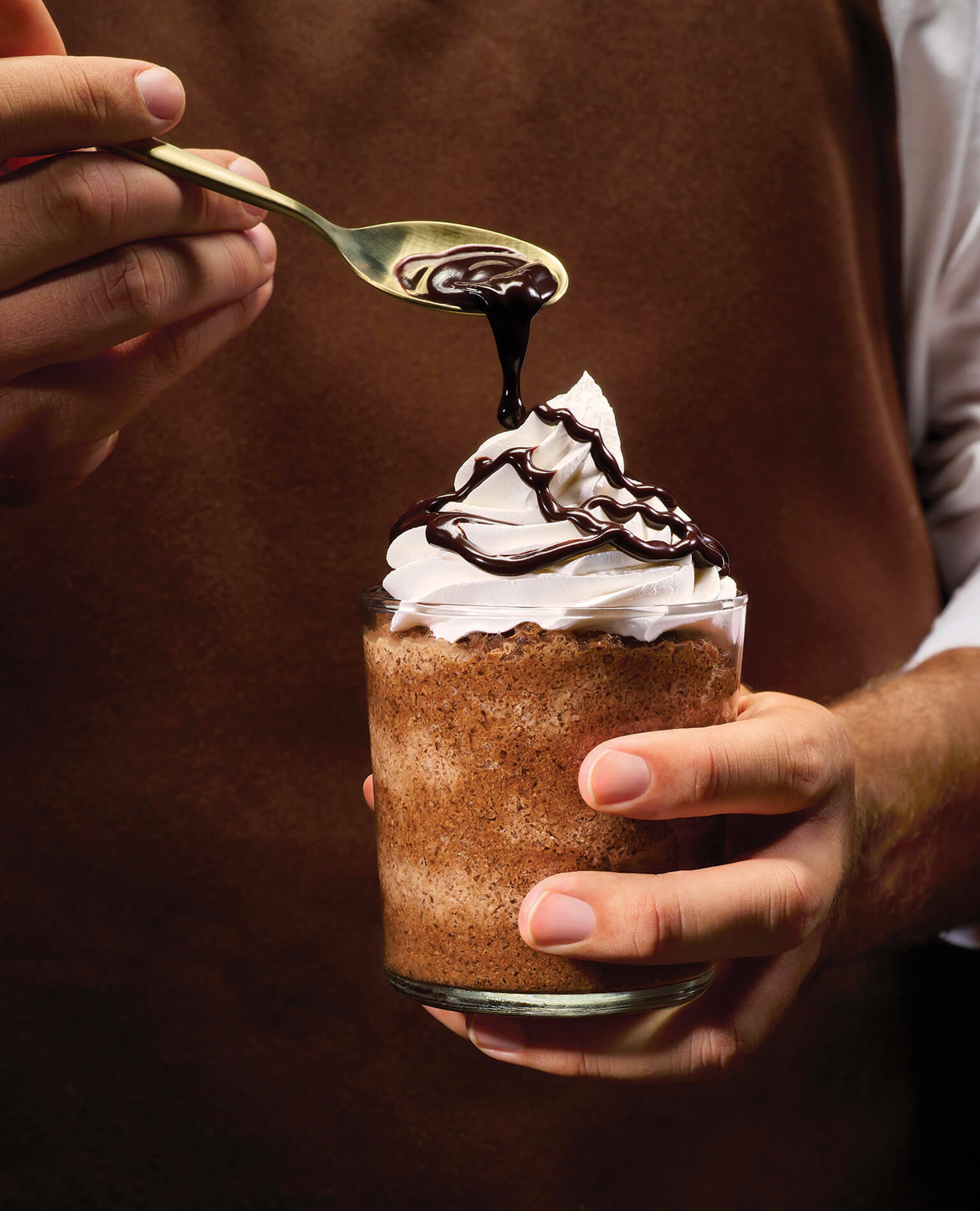 A man holding a coffee drink with a pile of whipped cream on top being drizzled with chocolate sauce.