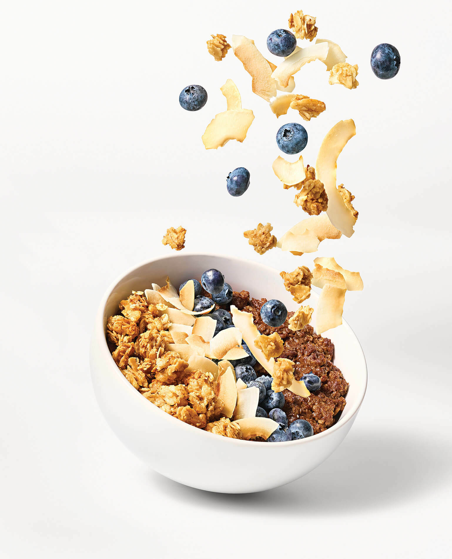 Blueberries, granola, and almond slivers falling into a bowl of oatmeal.