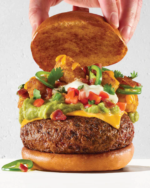 A hand placing the top bun on a large Loaded Tot-chos Topped Burger.