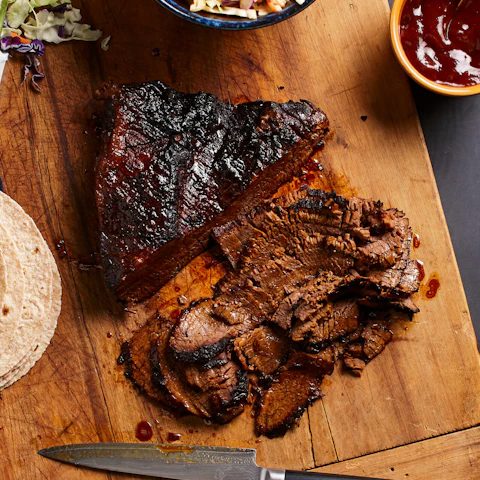 Sliced beef brisket on a cutting board next to a stack of tortillas, a small bowl of barbecue sauce, and a bowl of coleslaw.