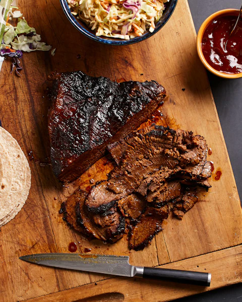 Sliced beef brisket on a cutting board next to a stack of tortillas, a small bowl of barbecue sauce, and a bowl of coleslaw.