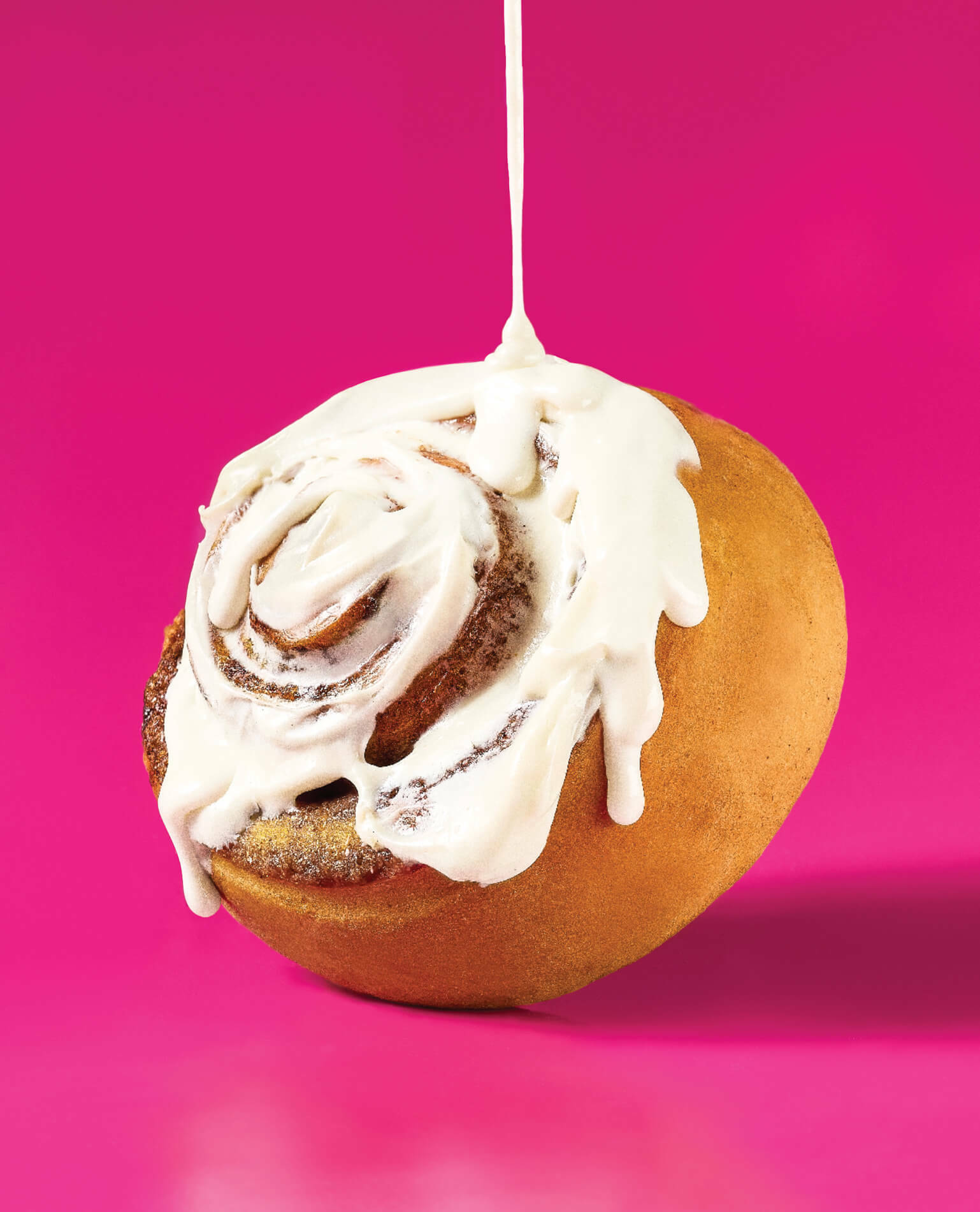 A cinnamon roll being drizzled with frosting on a pink background.