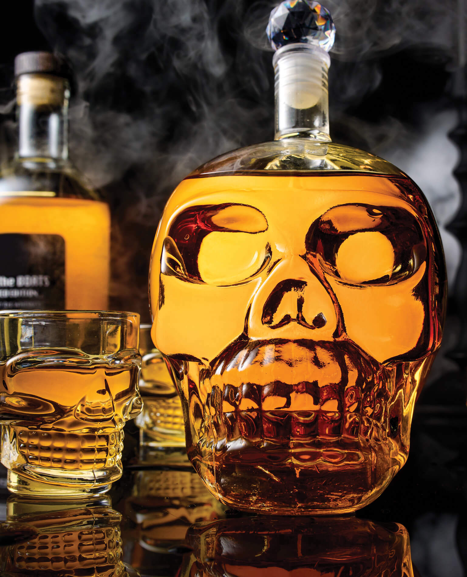 A spooky glass skull decanter with skull glasses on a smoky background.