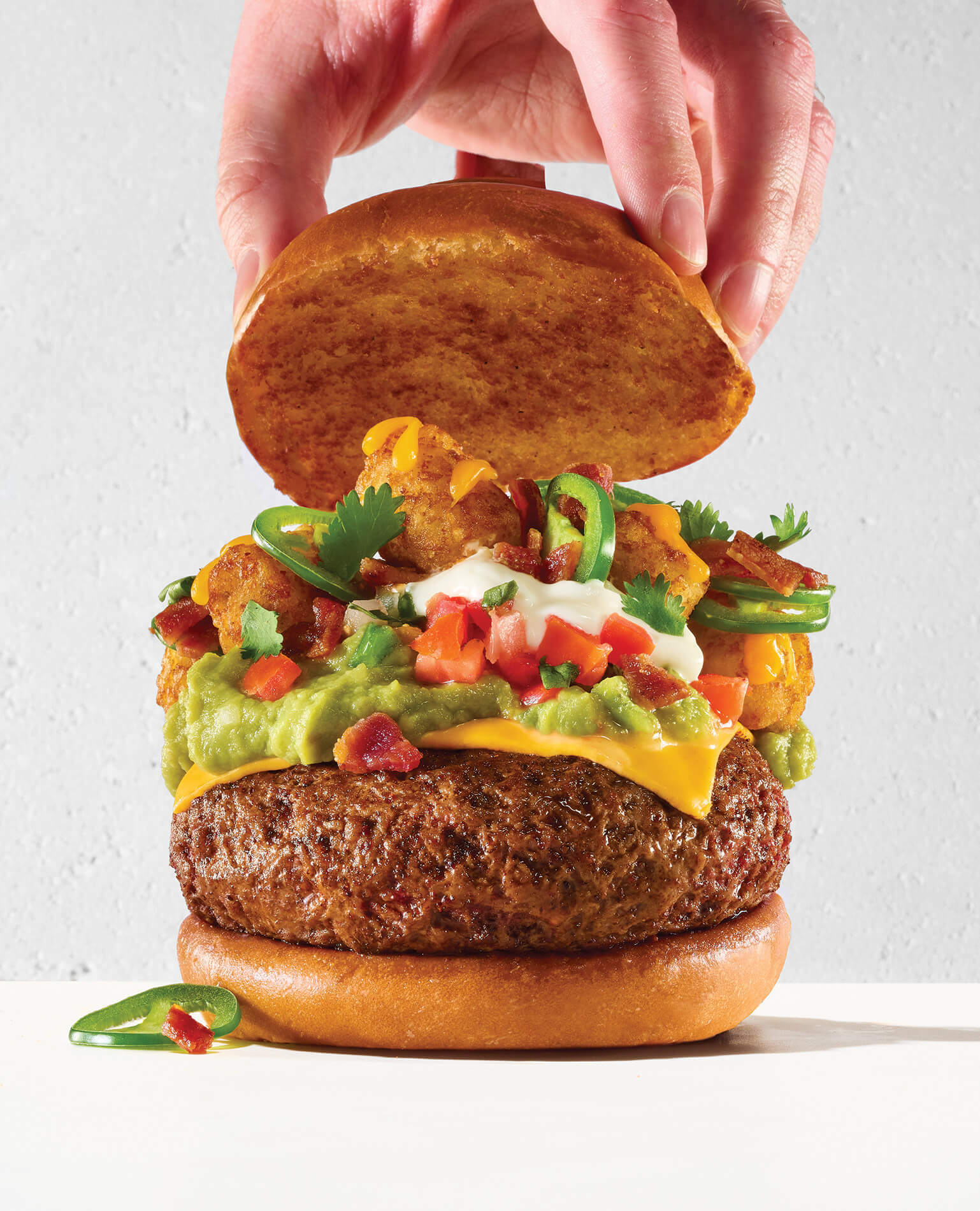 A thick cheeseburger stacked with guacamole and tater tots, with a hand lifting the top bun.