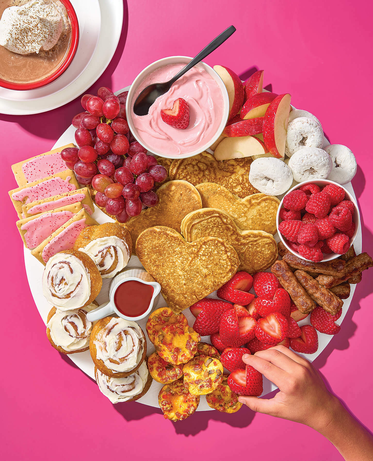 A child's hand reaching for a strawberry from a Valentine's Day breakfast charcuterie board.