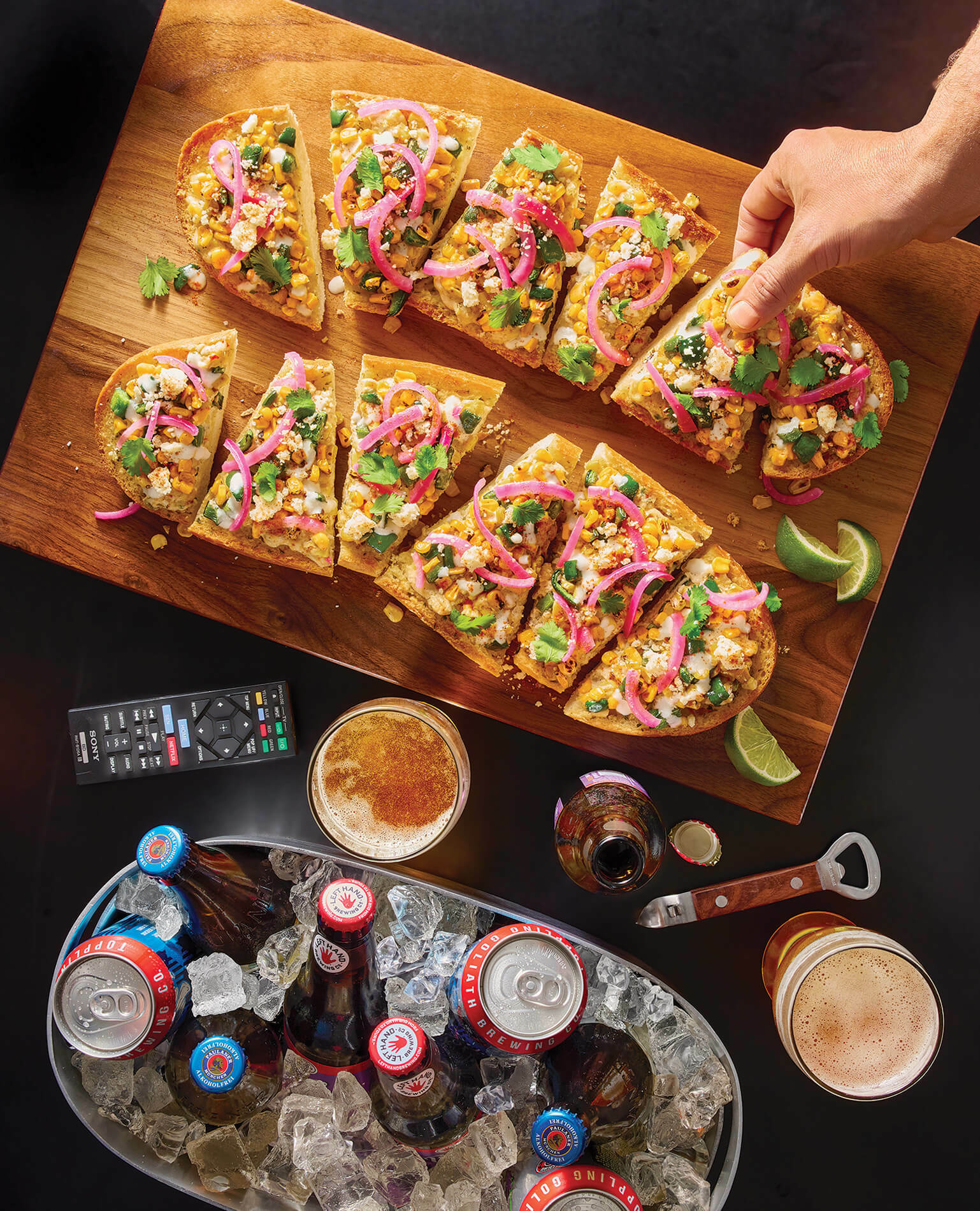 Hand reaching for a piece of garlic bread on a game watch party table complete with remote, cooler of drinks and glasses of beer.